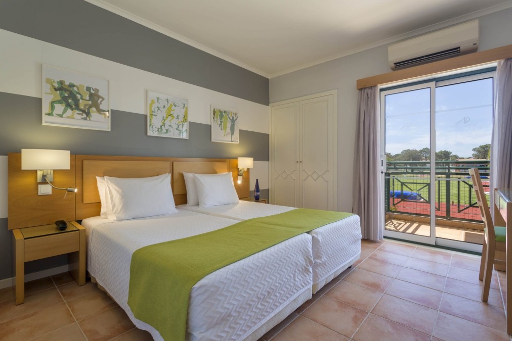 Chambre d'hôtes avec lits jumeaux, balcon et vue sur le terrain de sport.