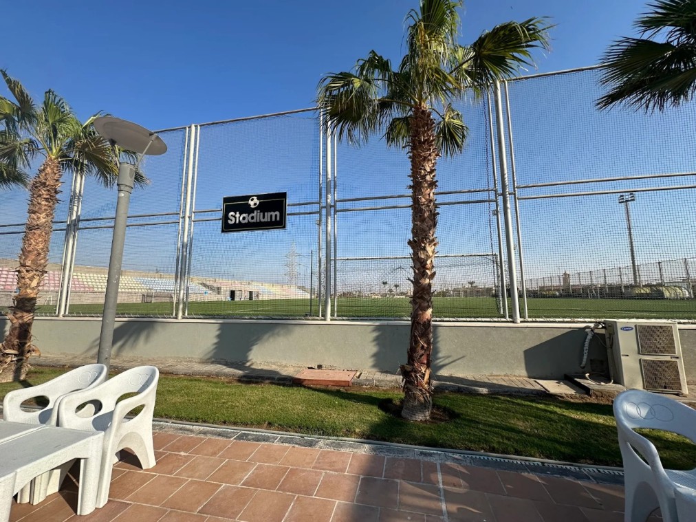 Vue du stade de football à travers un grillage métallique avec palmiers et chaises d'extérieur