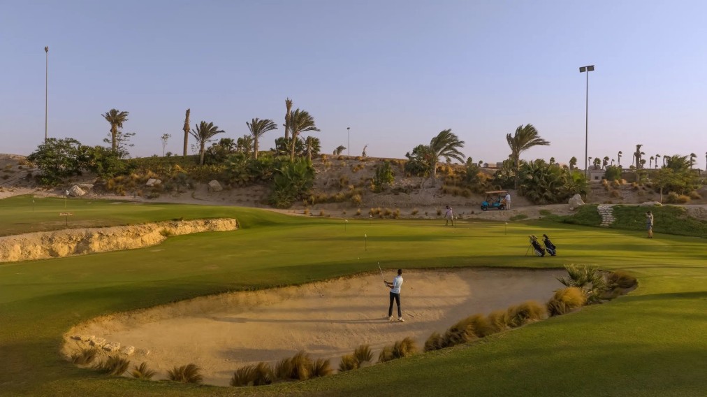 Golfeur jouant depuis un bunker sur un parcours verdoyant avec arbres et lumière du soir