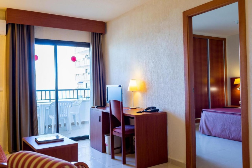 Espace de vie avec bureau, canapé et balcon, relié à une chambre.