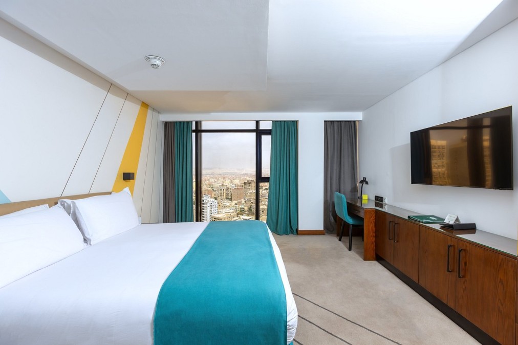 Chambre d'hôtel moderne avec lit blanc et chemin de lit turquoise, bureau et télévision, grande fenêtre donnant sur la ville