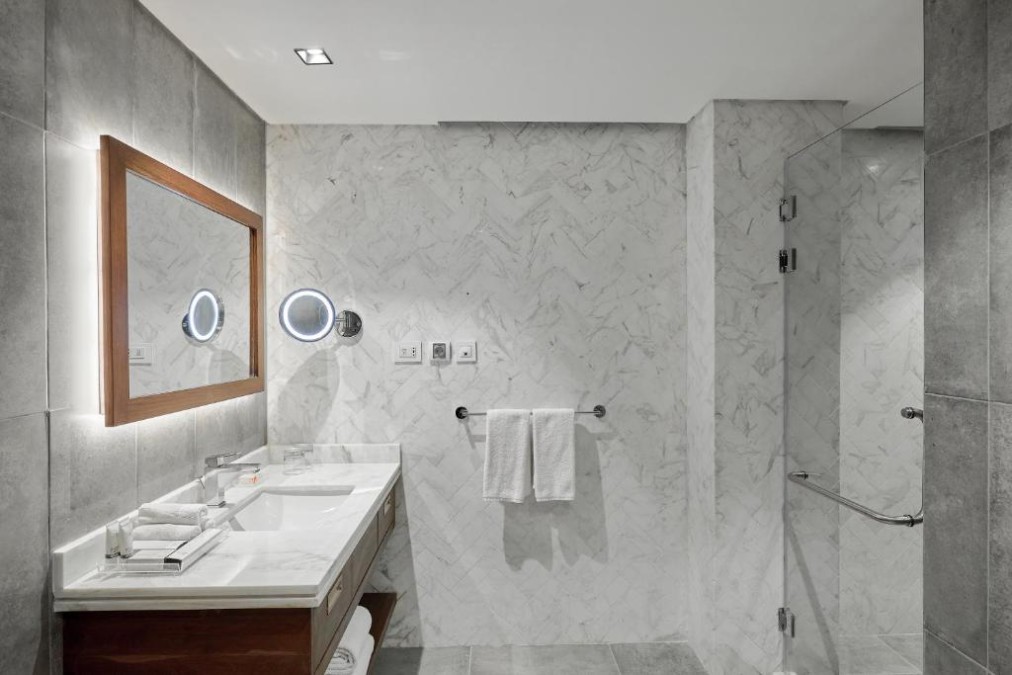 Salle de bain d'hôtel carrelée en gris avec murs en marbre, lavabo, miroir éclairé, porte-serviettes et douche vitrée